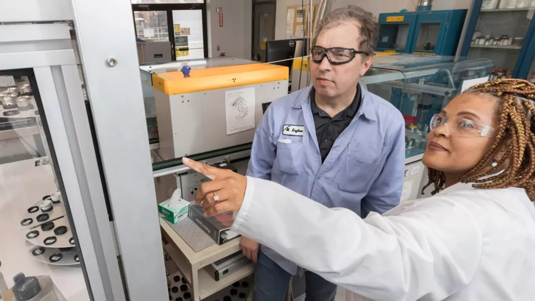 Two diverse scientists wearing lab coats and safety glasses are working in a laboratory. One scientist points at a piece of equipment while explaining something to the other. Various lab instruments and containers are visible in the background.