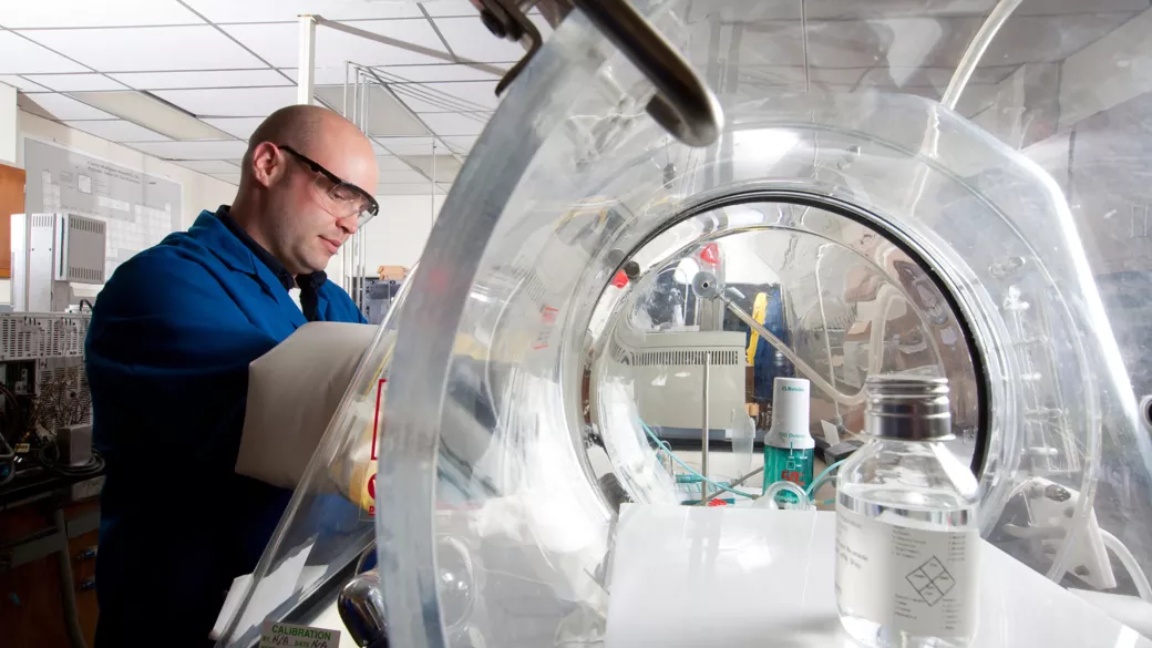 A person wearing safety glasses and a blue lab coat is working in a laboratory. They are using a glove box with a transparent dome to handle materials inside. Laboratory equipment and a large clear container are visible in the background.