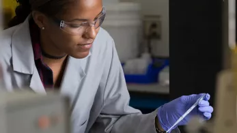 An Albemarle scientist wearing protective eyewear and gloves uses a pipette to add liquid to a small container in a laboratory. The scientist is focused on the task, and various lab equipment is visible in the background.