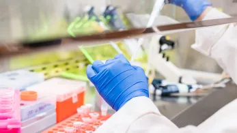 A scientist's gloved hand pipetting a sample in a lab filled with scientific equipment. Bright laboratory setting focused on experimentation and analysis.