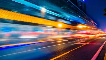 Long exposure shot of a busy city street at night showing streaks of lights from moving vehicles, with buildings illuminated by streetlights and signs.