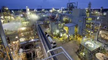 Aerial view of a large industrial complex filled with numerous interconnected metal structures, pipes, and tanks. The facility is illuminated with bright lights, indicating late evening or early morning operations. Smokestacks emit vapor, with the sky in a dusky hue.
