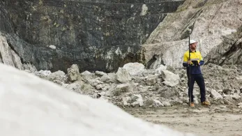 A lone worker in a yellow and blue uniform stands on rocky terrain at a quarry site, holding a surveying pole. The background features a steep rock face, with various shades of gray and brown indicating the layers of earth and mineral deposits.
