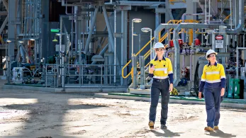 Two individuals wearing yellow safety jackets and white hard hats walk through an industrial facility. They are surrounded by machinery and pipelines. The weather appears sunny, and shadows fall on the ground, indicating bright sunlight.