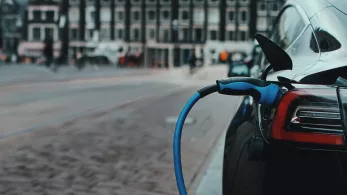 An electric car being charged on a city street, with the charging cable connected to the car's port and blurred buildings in the background.