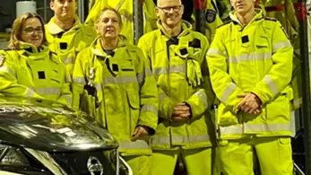 Five firefighters wearing high-visibility yellow gear stand together in a garage. Behind them are shelves with helmets and other firefighting equipment. A car with an Australian license plate is partially visible in the foreground.