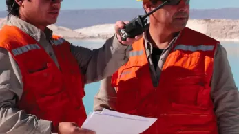 Two men wearing white and yellow hard hats and orange safety vests stand outdoors. One man holds a radio while the other holds a clipboard. They both wear sunglasses and are looking into the distance, with a body of water and mountains in the background.