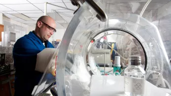 A person wearing safety glasses and a blue lab coat is working in a laboratory. They are using a glove box with a transparent dome to handle materials inside. Laboratory equipment and a large clear container are visible in the background.