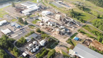 Aerial view of an industrial complex with large storage tanks, pipelines, and several buildings surrounded by grassy areas and trees. The facility features multiple storage tanks and an intricate network of structures and infrastructure.