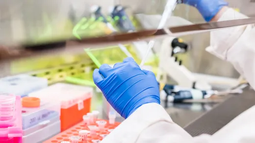A scientist's gloved hand pipetting a sample in a lab filled with scientific equipment. Bright laboratory setting focused on experimentation and analysis.