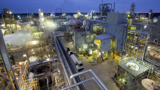 Aerial view of a large industrial complex filled with numerous interconnected metal structures, pipes, and tanks. The facility is illuminated with bright lights, indicating late evening or early morning operations. Smokestacks emit vapor, with the sky in a dusky hue.