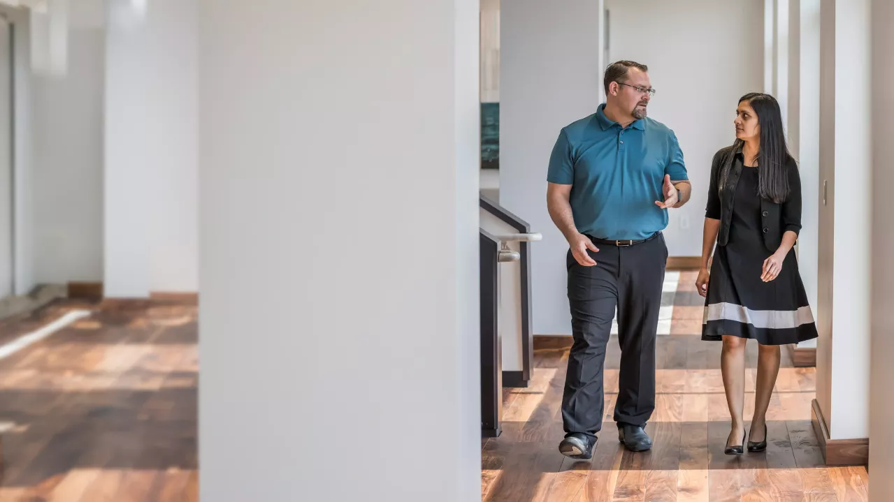 Two Albemarle employees walking and talking in an office corridor. Male wears a blue shirt and black pants, while the female wears a black dress. Office cubicles and dividers line the hallway they are walking through.