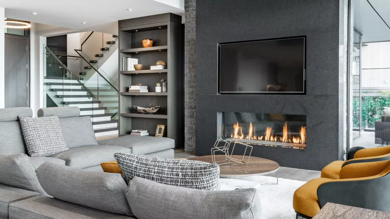 Modern living room with a large fireplace, gray sofas, patterned armchairs, and built-in shelves. Glass walls reveal a stairway and outdoor view.