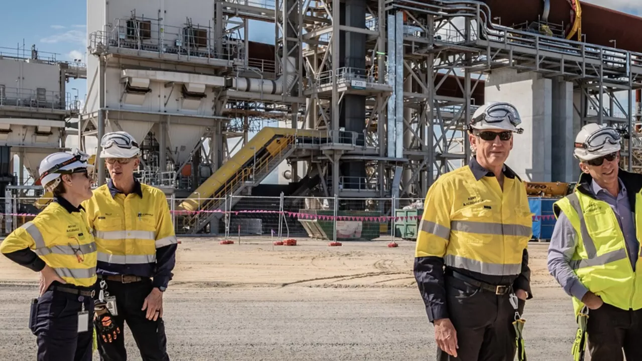 Kent Masters, Albemarle's CEO and his team in high-visibility clothing and hard hats discuss on a large industrial plant background, with heavy machinery and structures around.