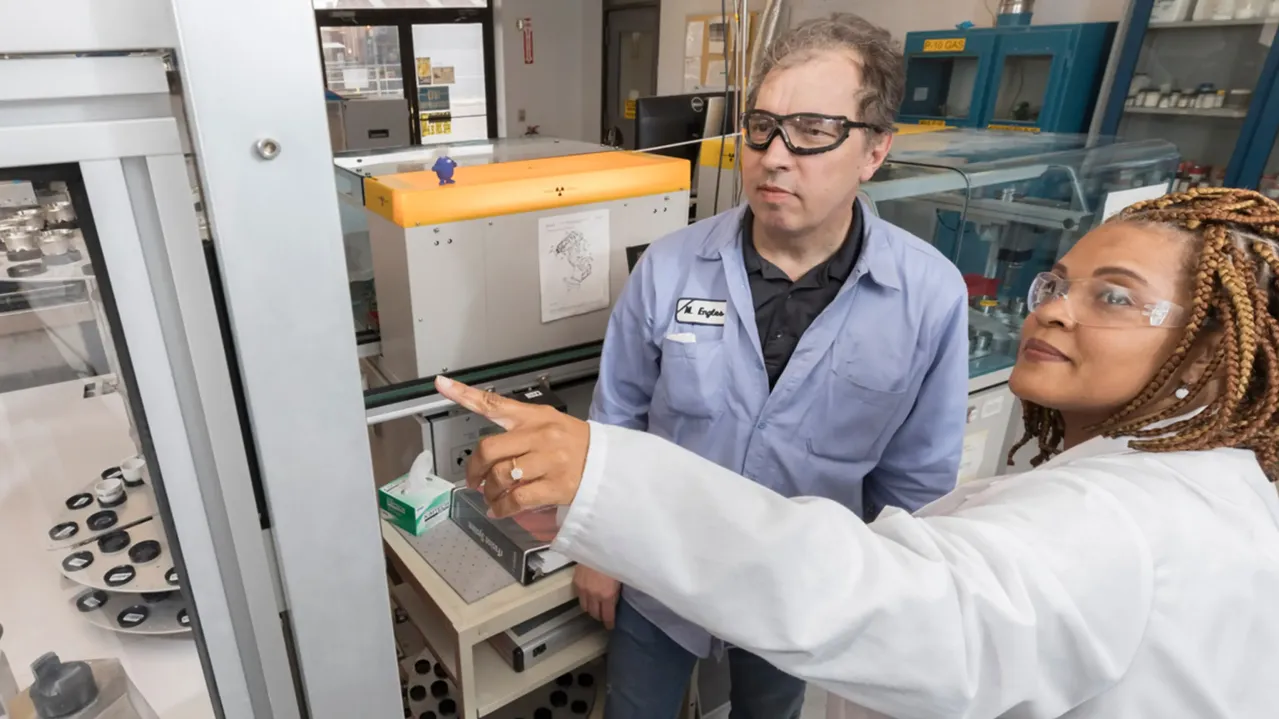 Two diverse scientists wearing lab coats and safety glasses are working in a laboratory. One scientist points at a piece of equipment while explaining something to the other. Various lab instruments and containers are visible in the background.