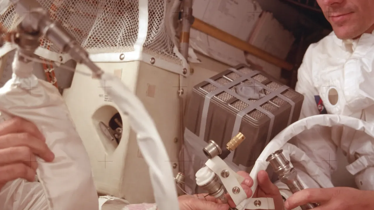 Astronaut Jack Swigert from the Apollo 13 mission in a space suit inside a spacecraft, closely examining the lithium hyrdoxide canister with another astronaut assisting, surrounded by equipment and cables.