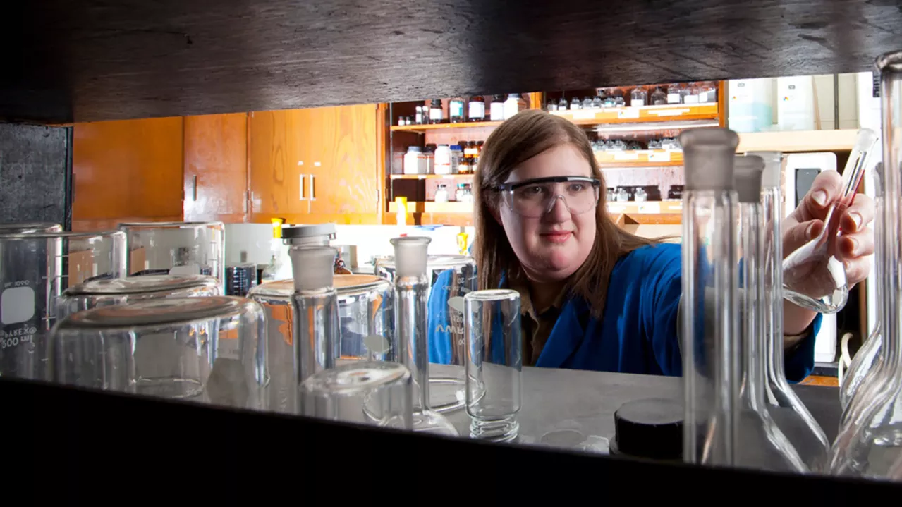 A person wearing safety goggles and a blue lab coat reaches for a glass beaker in a well-equipped laboratory. Various glassware and scientific equipment are arranged on a shelf in the foreground. Shelves with bottles and containers are visible in the background.