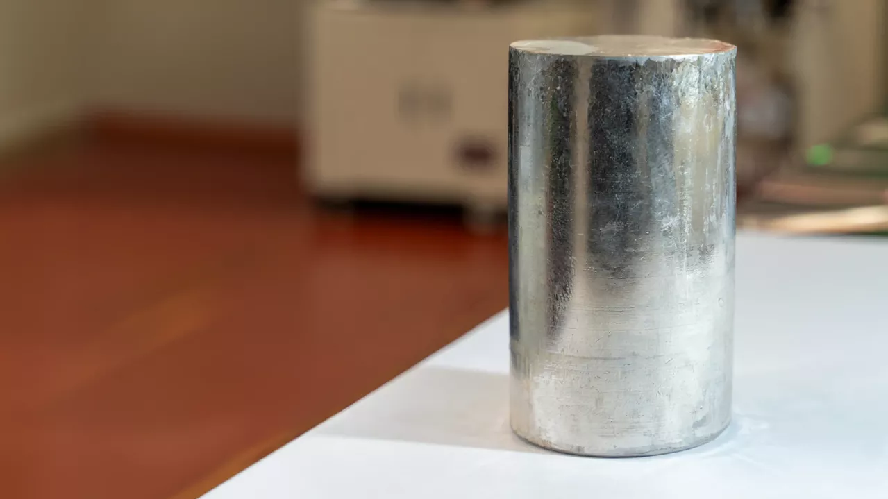 A cylindrical metal ingot on a white surface, exhibiting a slightly tarnished and dull surface, with blurred kitchen appliances in the background.