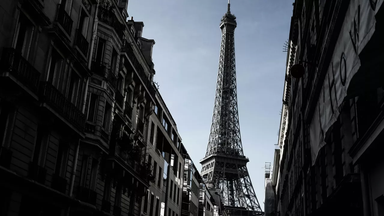 View from a narrow street in Paris flanked by traditional European buildings, looking towards the Eiffel Tower under a clear sky. The image captures the contrast between the shadowed street and the brightly lit tower.