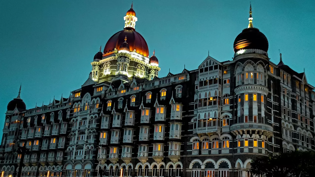 The Taj Mahal Palace Hotel illuminated at twilight, showcasing its iconic red domes and detailed Gothic architecture against a darkening sky.