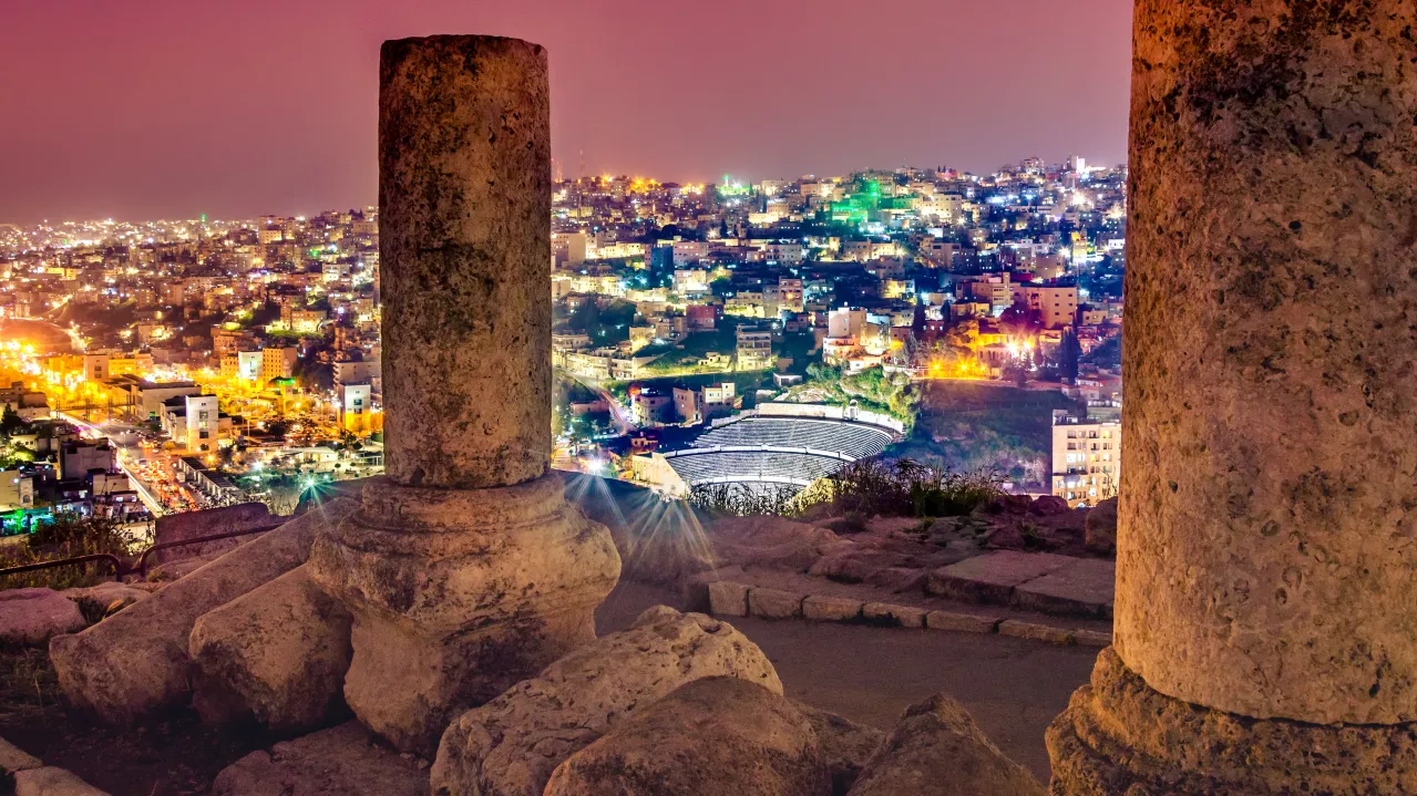The image shows a nighttime view of an amphitheater framed by two stone columns in the foreground, with a brightly lit cityscape sprawling out in the background. The city lights create a vibrant and lively atmosphere, contrasting with the historic ruins.
