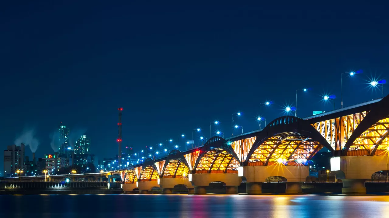 Night view of a lit-up bridge with multiple arches over a river, city skyline in the background, and a clear blue sky.
