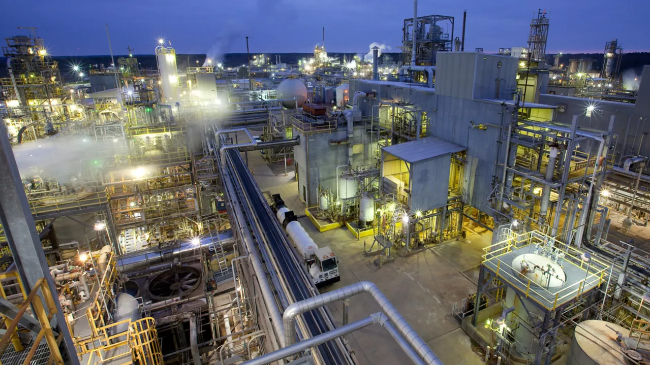 Aerial view of a large industrial complex filled with numerous interconnected metal structures, pipes, and tanks. The facility is illuminated with bright lights, indicating late evening or early morning operations. Smokestacks emit vapor, with the sky in a dusky hue.