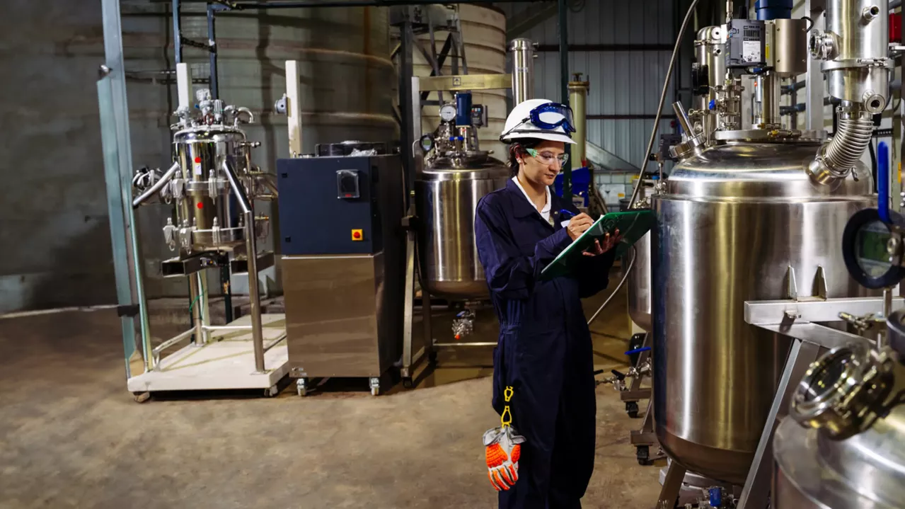 A worker wearing a hard hat and protective clothing stands in an industrial facility, writing on a clipboard. Large stainless steel tanks and various industrial equipment are visible in the background. The worker appears to be inspecting or documenting something.