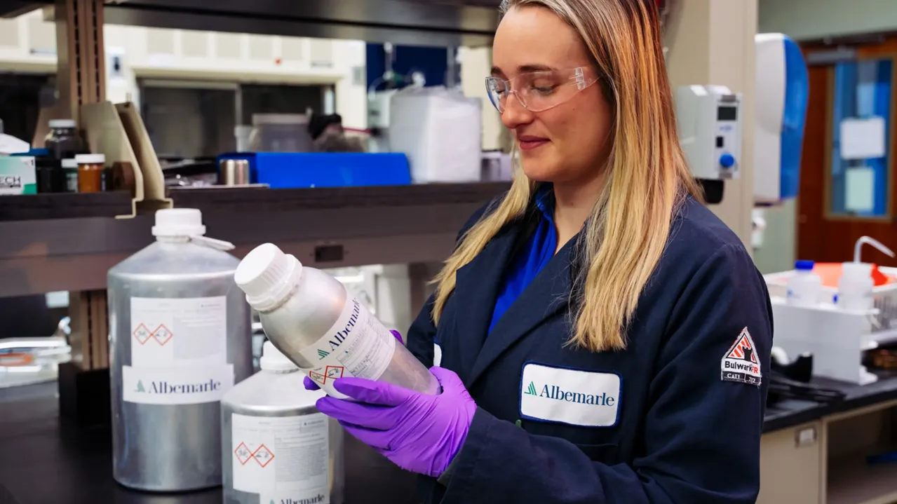 An Albemarle employee in a laboratory wearing safety goggles and purple gloves, holding a silver bottle with a label. They are dressed in a dark lab coat with a company logo. Shelves with various lab equipment and containers are visible in the background.