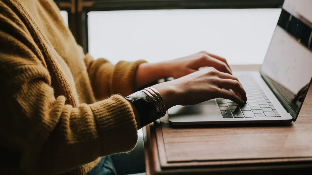 A person in a yellow sweater typing on a laptop with a strong, fire-resistant circuit board. The laptop is placed on a wooden desk, near a window with natural light streaming in.