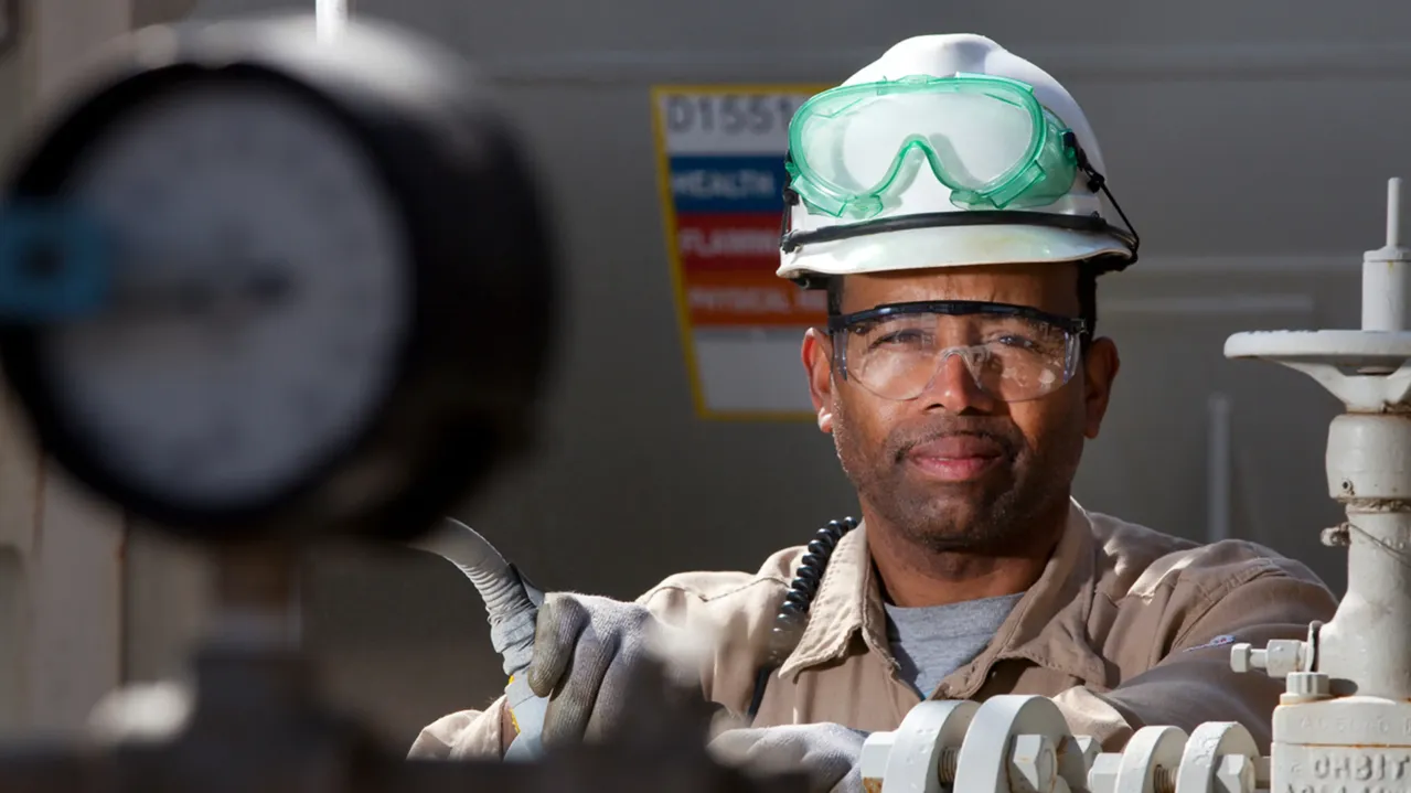A worker wearing a white hard hat, safety goggles, and gloves operates equipment in an industrial setting. He stands behind machinery, maintaining a focused expression, with other industrial components and a gauge partially visible in the foreground.