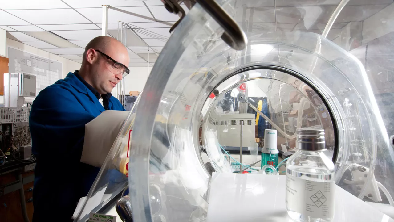 A person wearing safety glasses and a blue lab coat is working in a laboratory. They are using a glove box with a transparent dome to handle materials inside. Laboratory equipment and a large clear container are visible in the background.