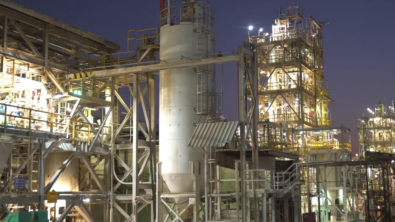 An industrial facility at dusk, featuring complex steel frameworks, pipes, and towers illuminated by artificial lights. The sky is a deep purple, indicating early nightfall. The image showcases the intricate and dense infrastructure of the plant.