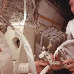 Astronaut Jack Swigert from the Apollo 13 mission in a space suit inside a spacecraft, closely examining the lithium hyrdoxide canister with another astronaut assisting, surrounded by equipment and cables.