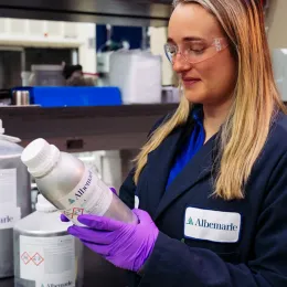 An Albemarle employee in a laboratory wearing safety goggles and purple gloves, holding a silver bottle with a label. They are dressed in a dark lab coat with a company logo. Shelves with various lab equipment and containers are visible in the background.