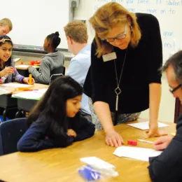 An elementary school classroom with a diverse group of students engaged in learning. A female teacher assists a young girl at a table, while other students work with adults in the background.