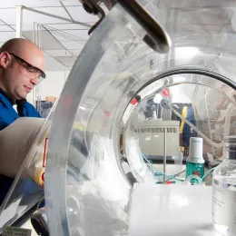 A person wearing safety glasses and a blue lab coat is working in a laboratory. They are using a glove box with a transparent dome to handle materials inside. Laboratory equipment and a large clear container are visible in the background.
