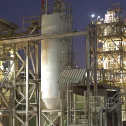 An industrial facility at dusk, featuring complex steel frameworks, pipes, and towers illuminated by artificial lights. The sky is a deep purple, indicating early nightfall. The image showcases the intricate and dense infrastructure of the plant.