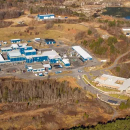 Aerial view of a large industrial facility with multiple blue-roofed buildings surrounded by trees and greenery. There is a smaller complex with white-roofed structures nearby. The area features paved roads, parking spaces, and two bodies of water in the background.