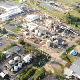 Aerial view of an industrial complex with large storage tanks, pipelines, and several buildings surrounded by grassy areas and trees. The facility features multiple storage tanks and an intricate network of structures and infrastructure.