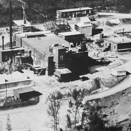 Historic black and white photo of an early 20th-century mining operation with various buildings, equipment, and earth mounds in a forested area.