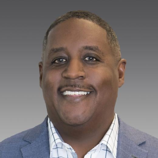 Headshot of Netha Johnson, President of Specialties, who is an African American man smiling with short black hair, wearing a dark blue suit with a white and blue striped shirt, against a grey background