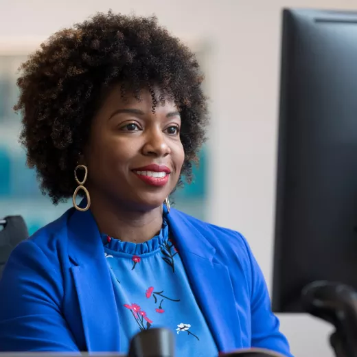 A woman with curly hair, wearing a blue blazer over a floral blouse, smiles while sitting at a desk. She has statement earrings and appears to be working on a computer. The background is blurred, focusing attention on her cheerful expression.