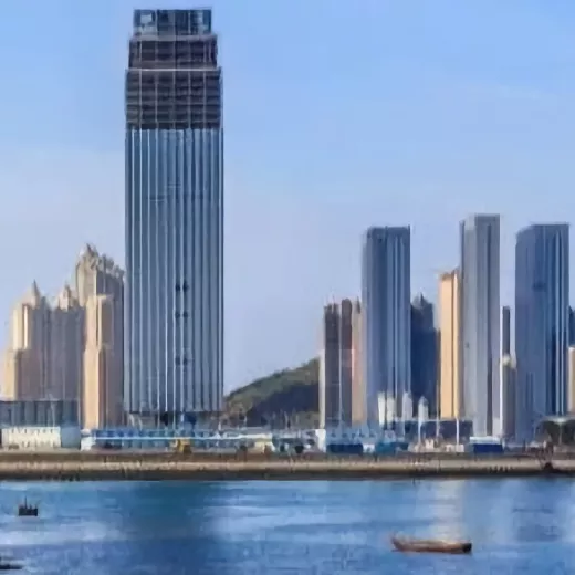 Tall modern skyscrapers in Dalian China with a prominent, unfinished high-rise building stand along a waterfront, reflecting in the calm blue water. A small boat is visible in the foreground, and a green hill is in the background under a clear sky.