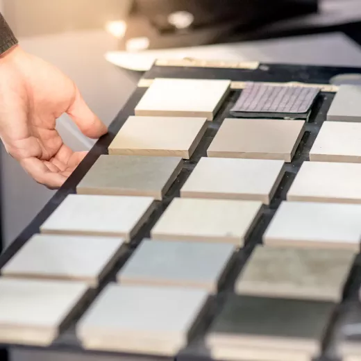 A person's hand near a display of various tile samples, arranged neatly on a table, in a home improvement store. The focus is on the tiles, with a soft background.