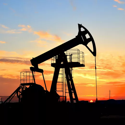 Silhouette of an oil pump jack operating at sunset with a vibrant orange sky in the background.