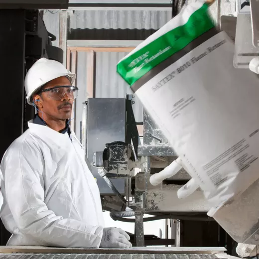 A worker wearing a white hard hat, safety glasses, ear protection, white gloves, and a white protective suit stands next to industrial machinery. In the foreground, a bag labeled "SAYTEX 8010 Flame Retardant" is being moved by the machinery.