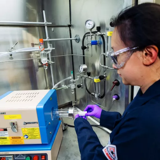 A scientist in safety goggles and gloves operates a sophisticated laboratory device with various dials and tubes, focusing intently on her task.