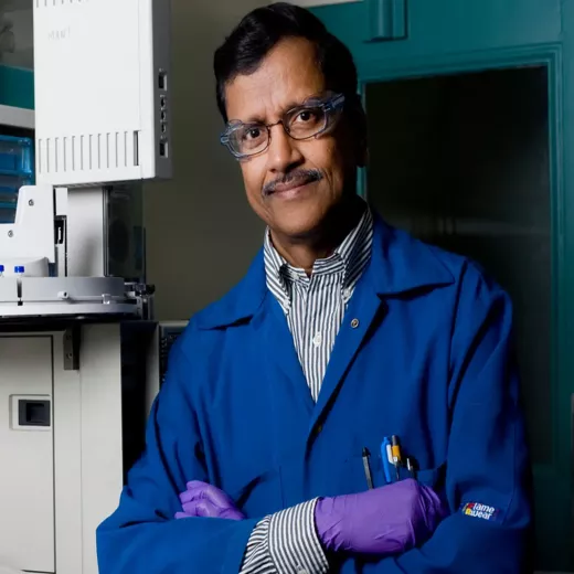 A middle-aged man wearing a blue lab coat, safety glasses, and purple gloves stands confidently beside a laboratory machine, with pens in his coat pocket.