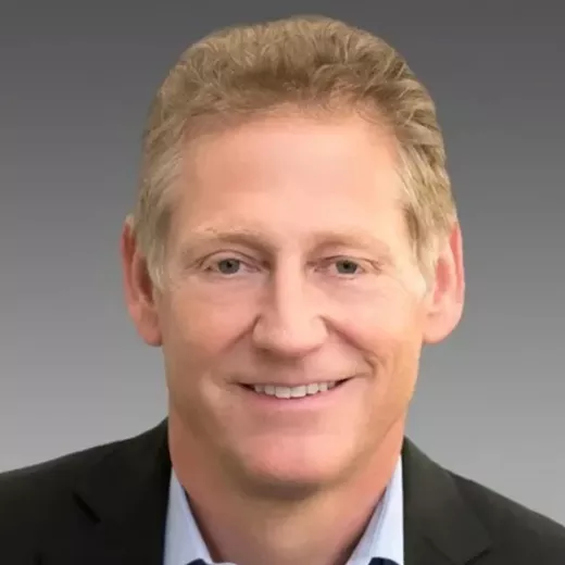 Headshot of Kent Masters, CEO of Albemarle a smiling Caucasian man with light blonde hair, wearing a dark suit and a white shirt, against a gray background.